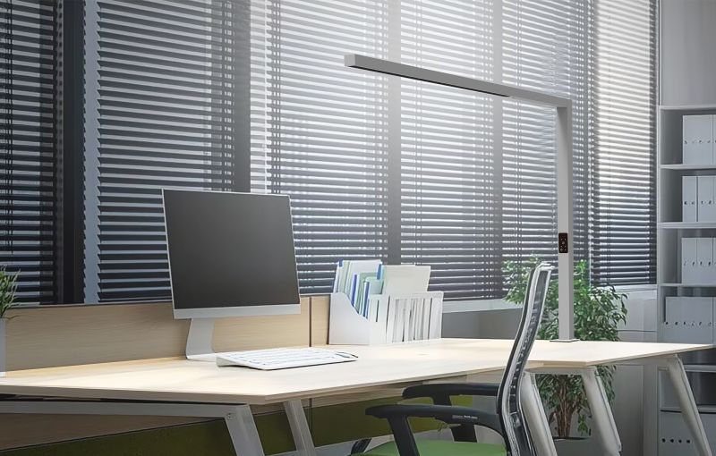 A linear luminaire beside a monitor at a desk with window blinds